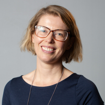 Smiling woman with short blonde hair, glasses, and a navy top against a plain grey background.